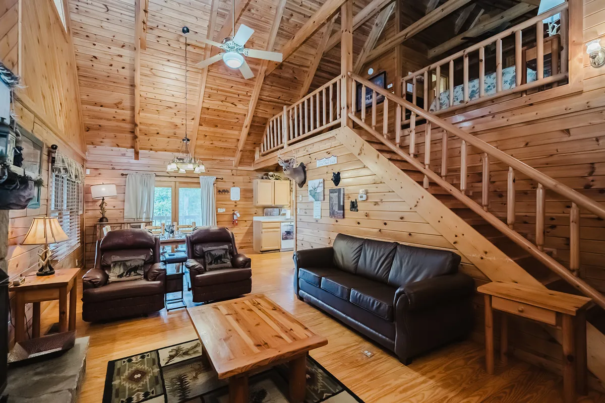 A spacious living room featuring exposed wooden beams, plush leather sofas, and a large wooden coffee table. Expansive windows fill the space with natural light, creating a seamless connection to the outdoors. The inviting arrangement encourages group relaxation and socializing after adventurous days.