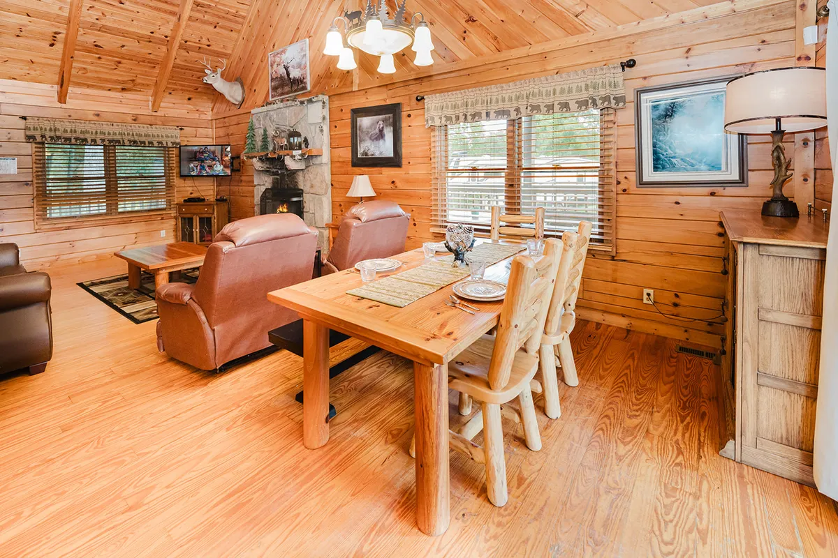 A cozy, open-concept living and dining area with a wooden dining table, comfortable armchairs, a fireplace, decorative artwork, and large windows. The space is perfect for family meals, game nights, and relaxation.
