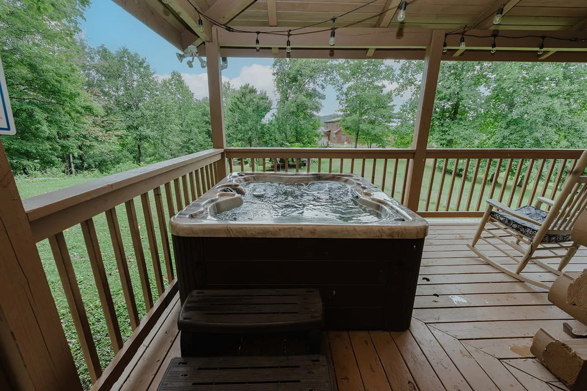 This image features a wooden deck nestled amidst lush greenery, with a bubbling hot tub as its centerpiece. The scene evokes tranquility, with the hot tub inviting guests to unwind while enjoying scenic mountain views and the peaceful natural surroundings. Subtle lighting hints at the possibility of romantic evenings under the stars.