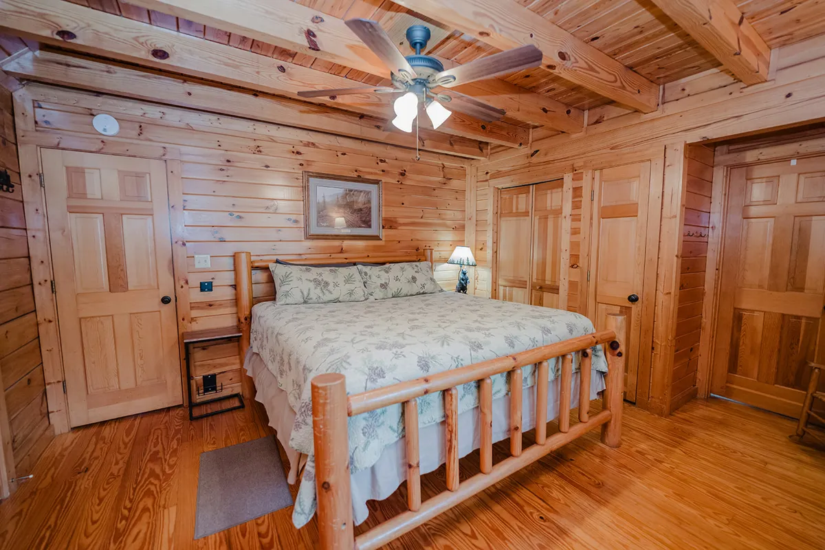 A cozy bedroom with a wooden bed frame and floral bedspread, accompanied by a nightstand with a lamp and wooden walls. The ceiling fan and soft lighting create an intimate, tranquil setting ideal for rest and reading.