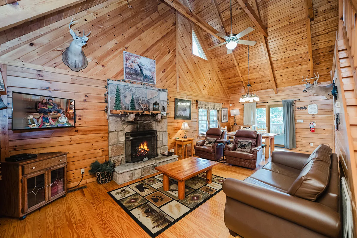 A warm, rustic living room with exposed wooden beams, a glowing fireplace at its center, plush seating including leather sofas and recliners, and artwork adorning the walls. The layout is open and inviting, suggesting plenty of space for family gatherings, relaxation, or movie nights. Light from the fire and surrounding lamps gives the space a cozy, welcoming glow.