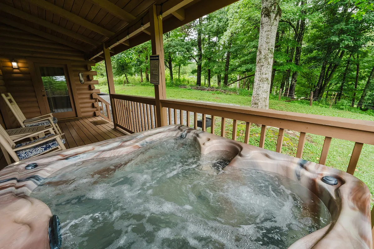 A wide wooden deck nestled among the Smoky woods, featuring a bubbling hot tub with a view of the surrounding trees. Rocking chairs are set along the railings, and the fading light of sunset creates a tranquil, inviting atmosphere.