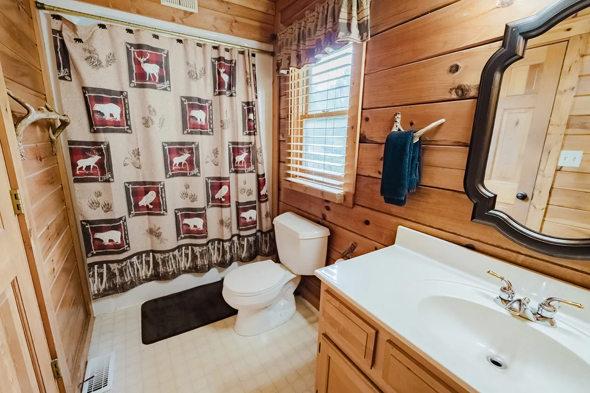 An inviting bathroom with wooden walls, a decorative shower curtain, and a well-lit vanity area with a mirror. The natural materials and design elements evoke warmth and comfort, ideal for unwinding after a day in nature or preparing for restful sleep.