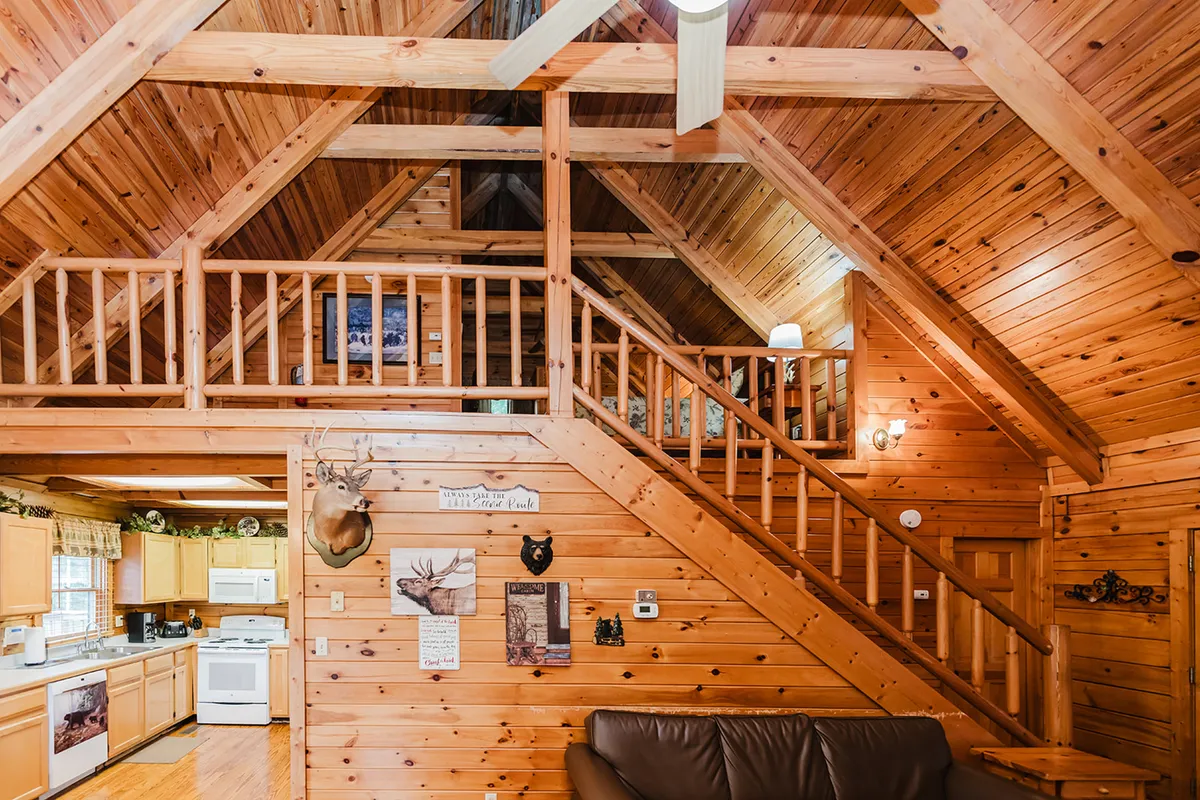 An open-plan interior displays wooden beams, an open staircase, a modern kitchen area with visible appliances, and a comfortable sofa nearby. The scene captures the flow between kitchen, dining, and living spaces, suggesting a warm environment for both meal prep and socializing.