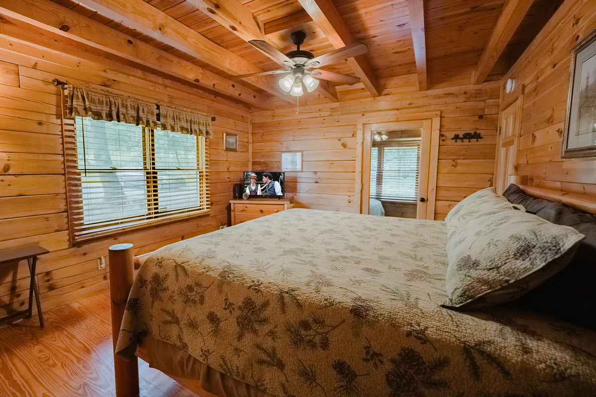 This image features a spacious bedroom anchored by a king-sized bed with a wooden frame, complemented by large windows that let in an abundance of natural light. The room also showcases a TV unit and inviting seating, all set against warm wooden walls and ceiling. A ceiling fan and plush linens create a comfortable, restful atmosphere.