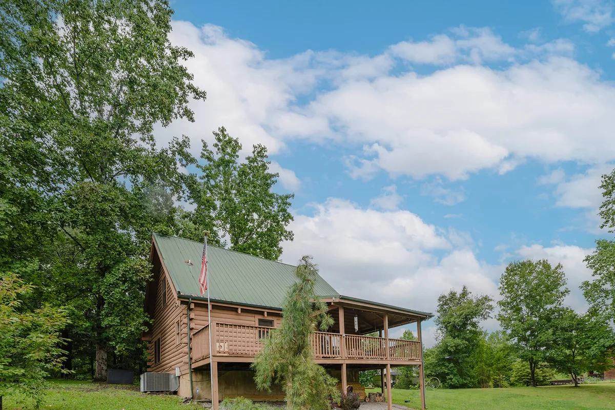 This image showcases a classic log cabin structure with a green metal roof nestled among lush trees. The spacious front porch, adorned with an American flag, offers a welcoming atmosphere for families. The surrounding natural setting hints at both tranquility and adventure, with open spaces for kids to play and families to gather.