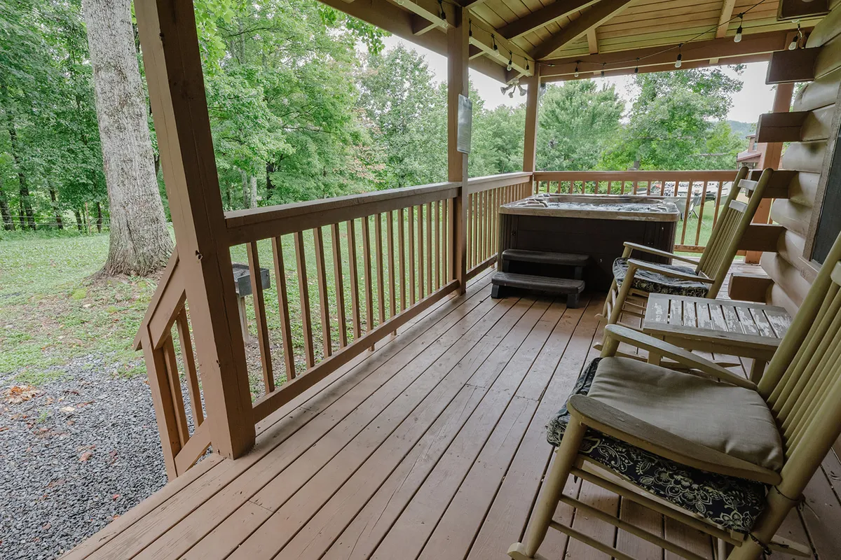 This exterior scene captures a peaceful deck area with rocking chairs, a hot tub, wooden railing, and a view of rolling green landscape. The setup invites relaxation and conversation, with nature views that reinforce the cabin’s proximity to outdoor adventures.