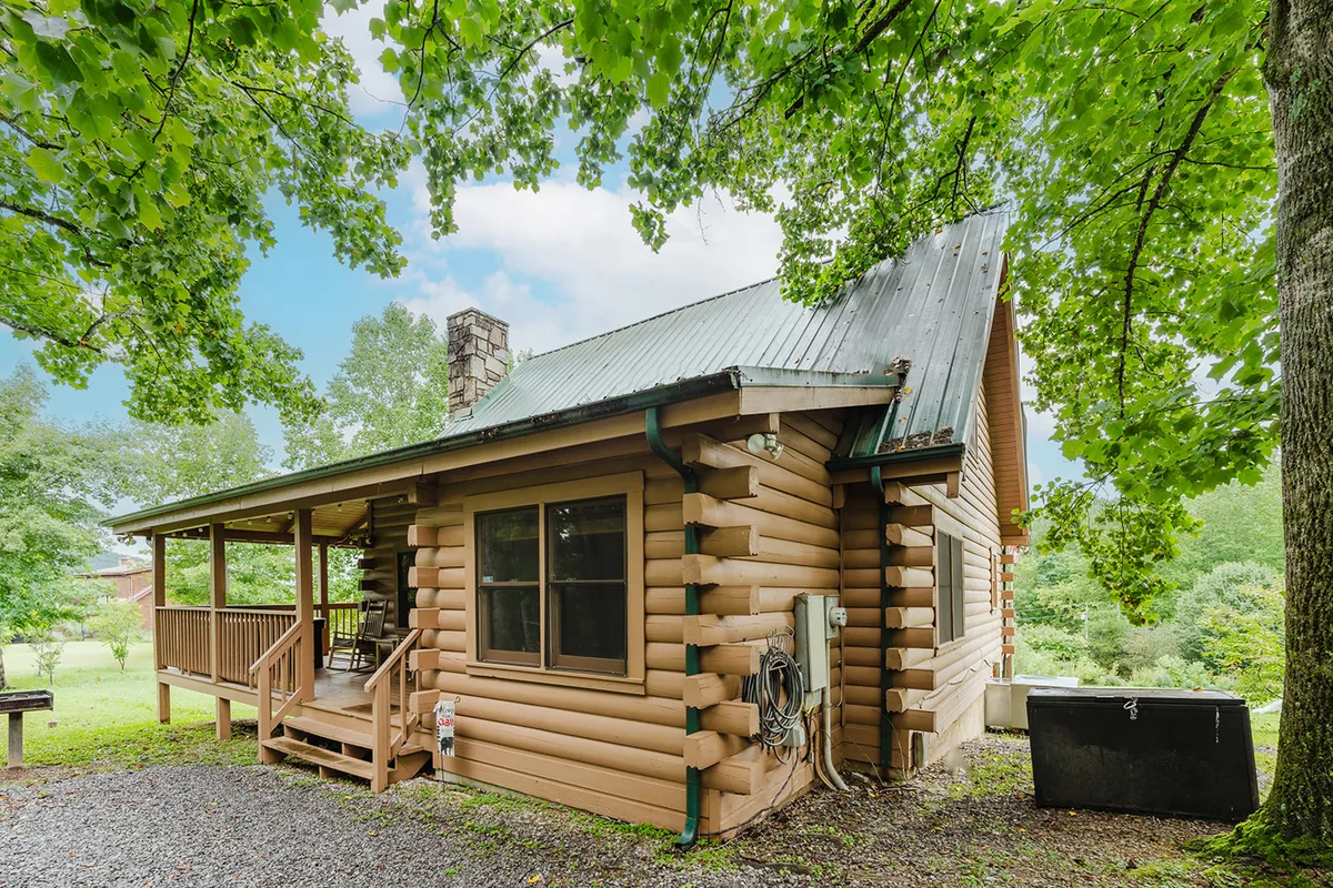 A classic log cabin exterior surrounded by lush green foliage, with a spacious porch area ideal for relaxing in the shade or gathering with friends and family. The inviting porch overlooks the natural setting, blending seamlessly into the tranquil mountain landscape.