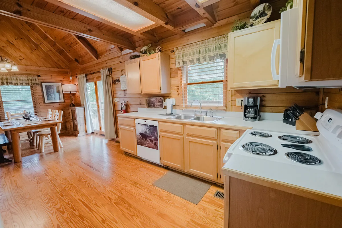 This image features a bright, inviting kitchen and dining space with a sturdy wooden countertop, a dining table set for four, and exposed wooden beams. Large windows flood the area with natural light, highlighting the seamless connection between kitchen and dining zones, perfect for preparing and sharing meals together.